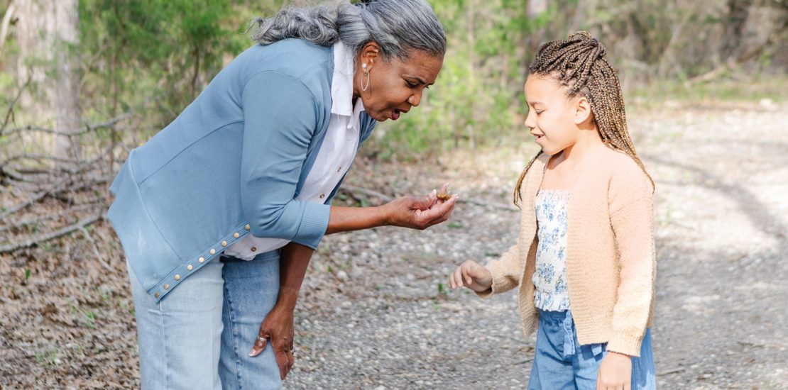 Enjoying a nature hike together, a grandmother leans in to show her granddaughter something she found on their path
